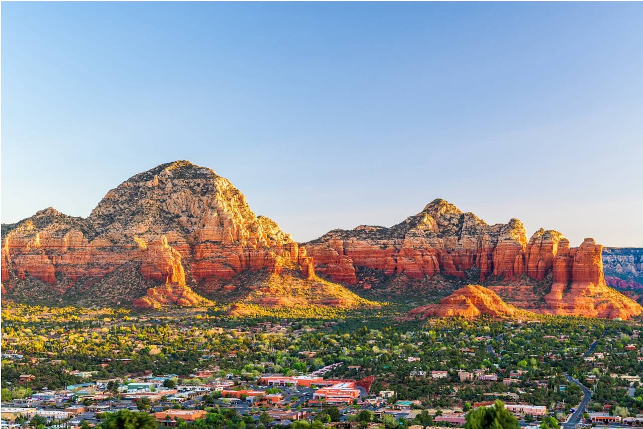 Panoramic view of Sedona, Arizona, with red rock formations and clear blue sky. A town with scattered buildings lies in the foreground.